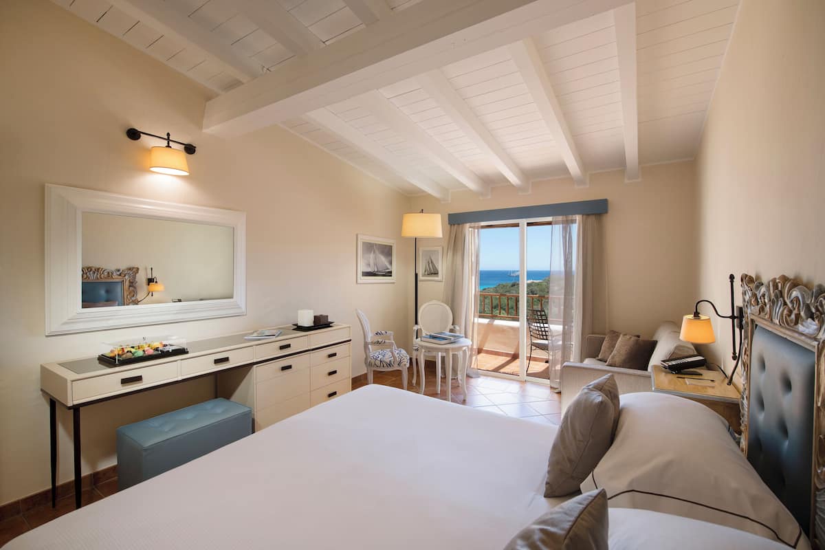 A hotel room with a large bed, a desk with drawers, a chair, a small table, a lamp, and a view of the sea through a sliding glass door.