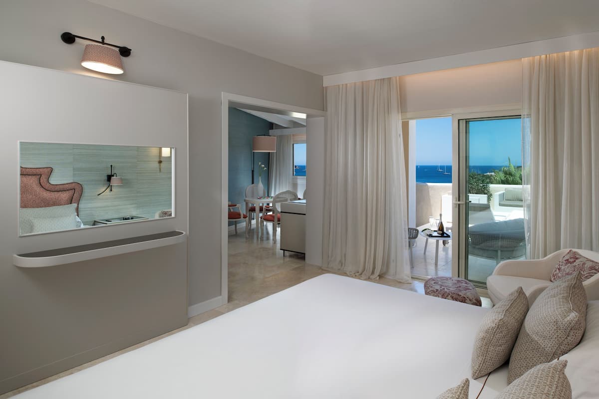 A modern hotel room with a large bed, a sitting area with a view of the sea, and a dining area.