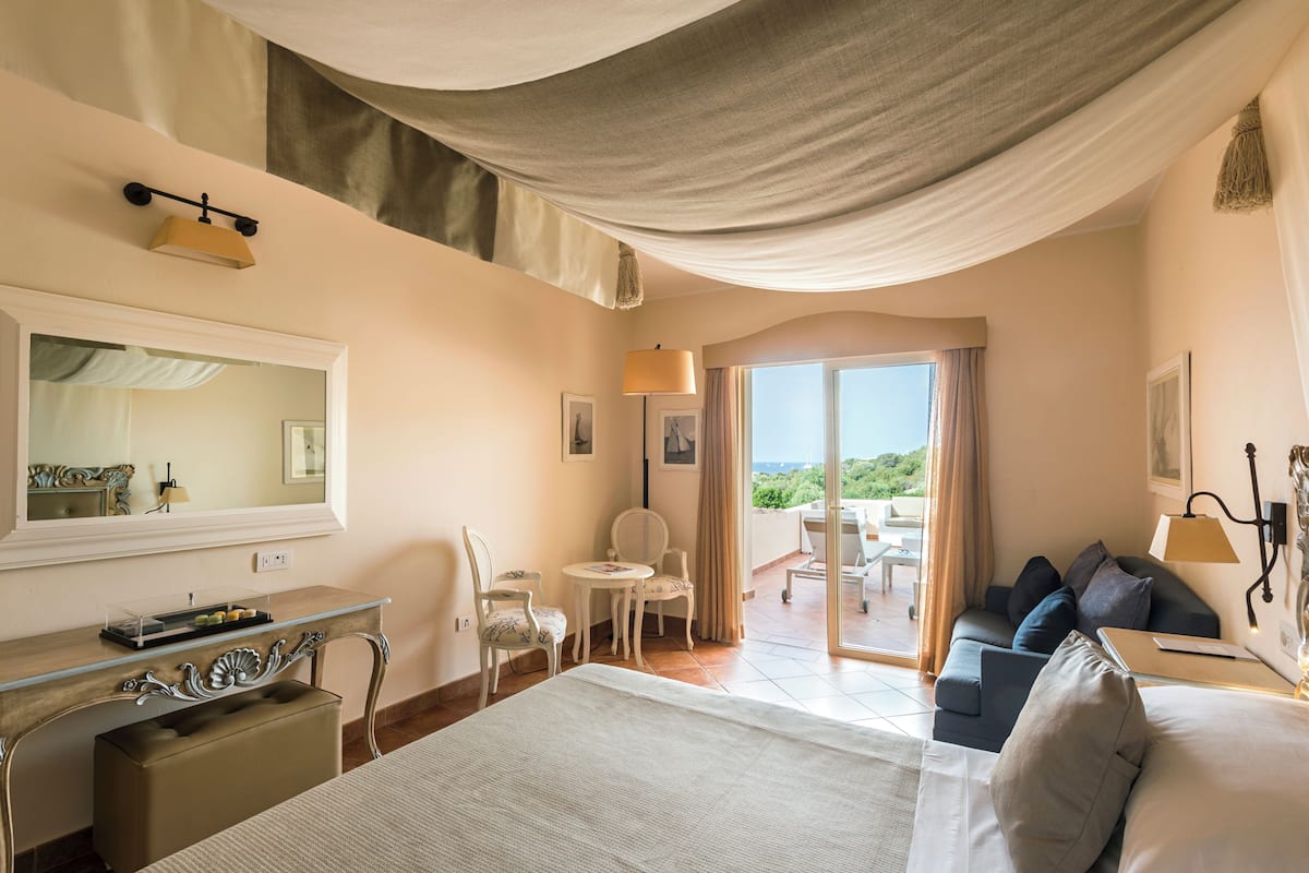 A hotel room with a bed, a desk, a chair, a sofa, a balcony with a view, and a large window.