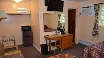 A hotel room with a bed, a desk with a chair, a television mounted on the wall, a small refrigerator, a trash bin, and a heater.