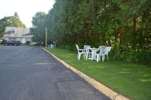 Una calle residencial con una casa, un coche aparcado y mobiliario de jardín en el césped.