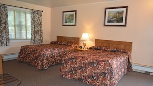 Quarto de hotel com duas camas, uma mesa de cabeceira com abajur, uma janela com cortinas e duas fotos emolduradas na parede.