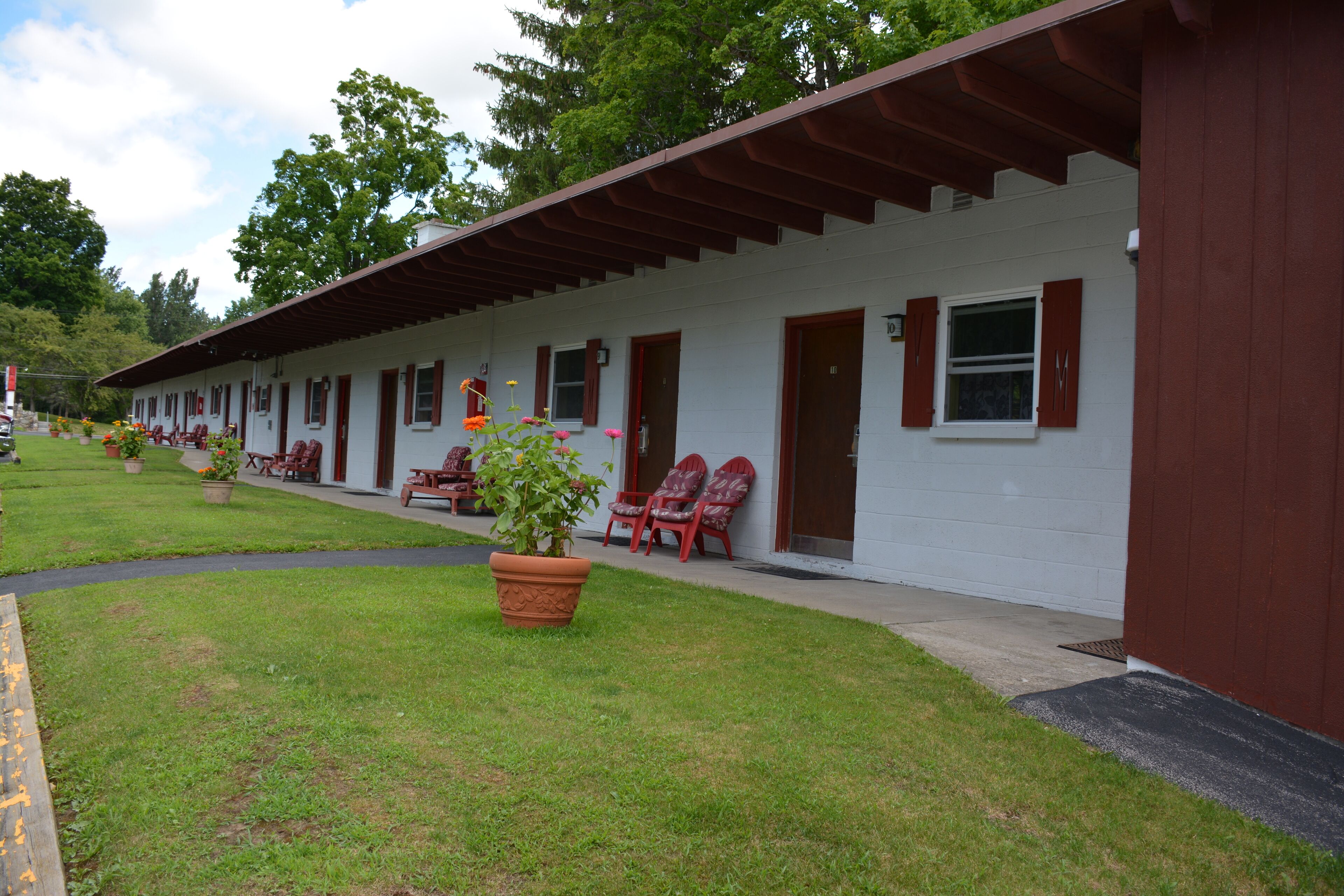 Un motel con una hilera de habitaciones, cada una con puerta y ventana, y un pequeño porche con sillas.