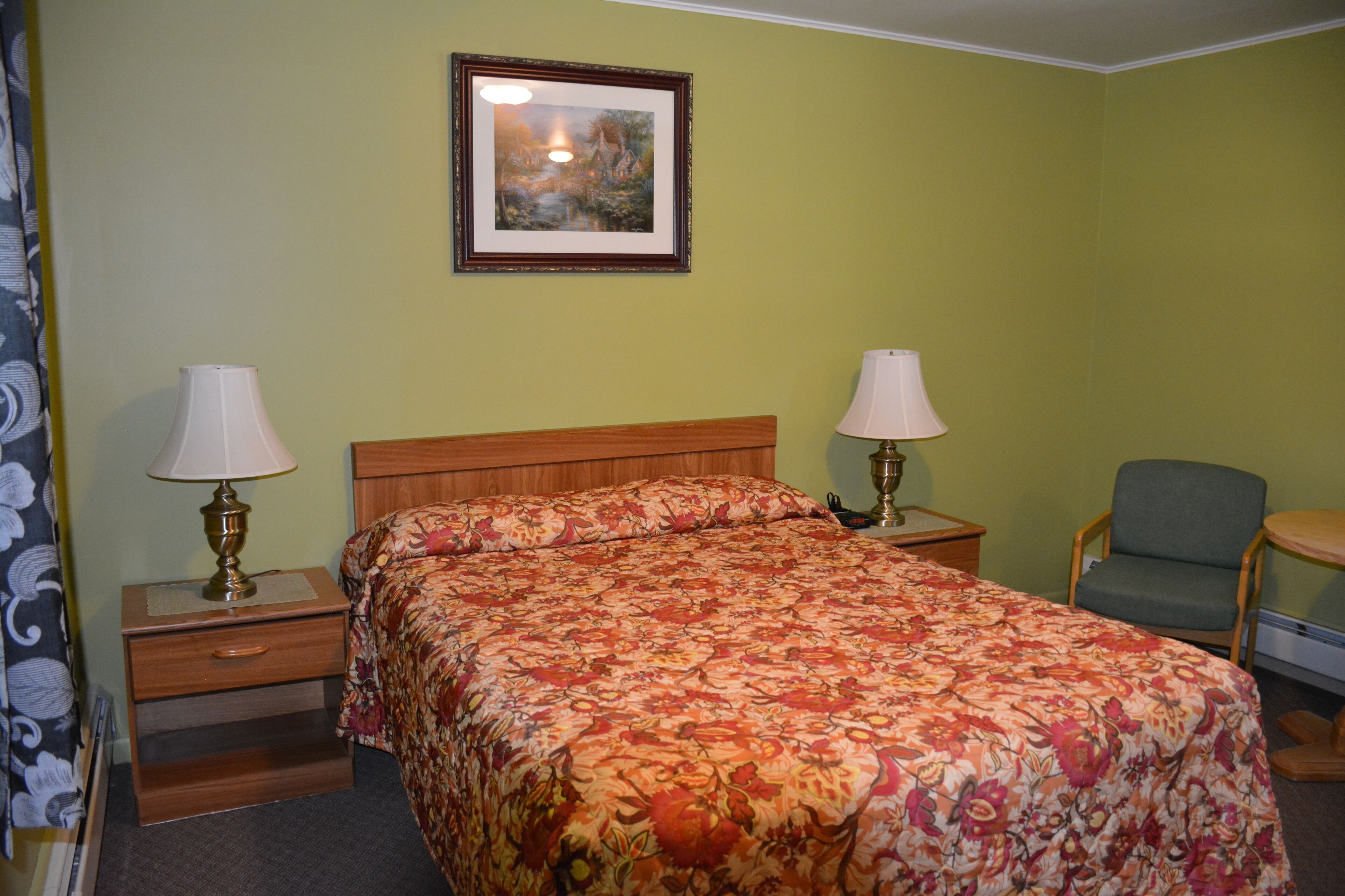 A hotel room with a floral bedspread, two bedside lamps, a wooden nightstand, a chair, and a framed picture on the wall.