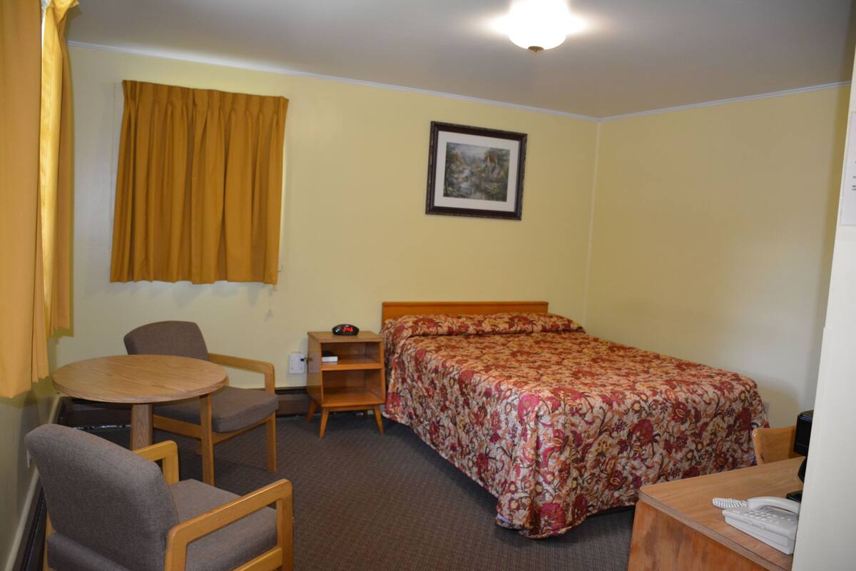 A hotel room with a bed, a desk, a chair, a small table, a nightstand, and a picture on the wall.