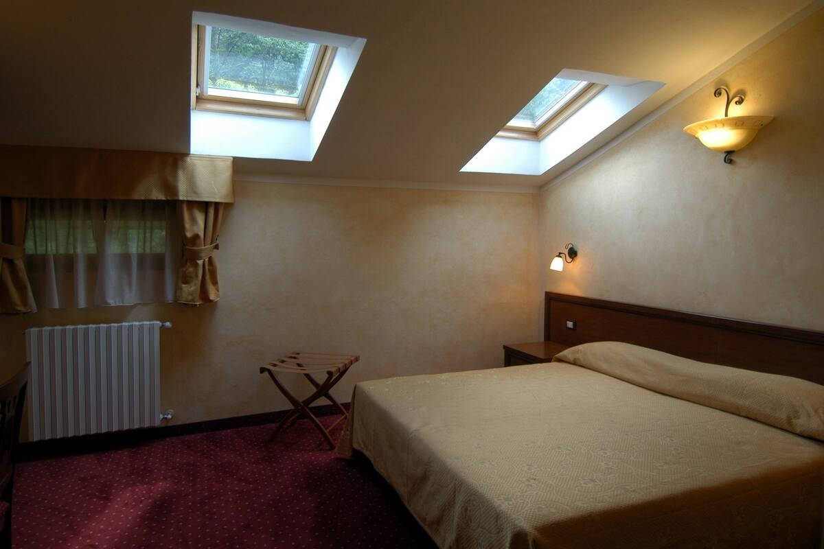 Un dormitorio con dos tragaluces, una cama, una mesita de noche, una silla y un radiador.