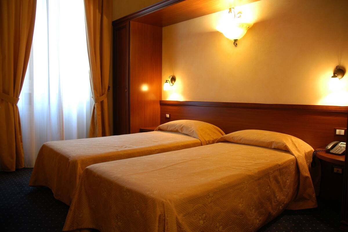 Habitación de hotel con dos camas, cabecero de madera, mesita de noche con teléfono e iluminación en la pared.
