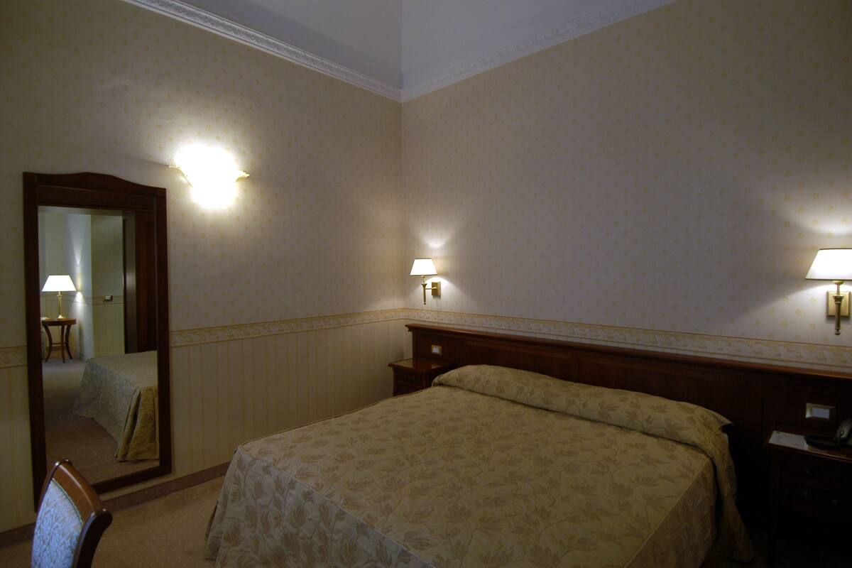 Habitación de hotel con cama, mesitas de noche, un espejo y lámparas de pared.