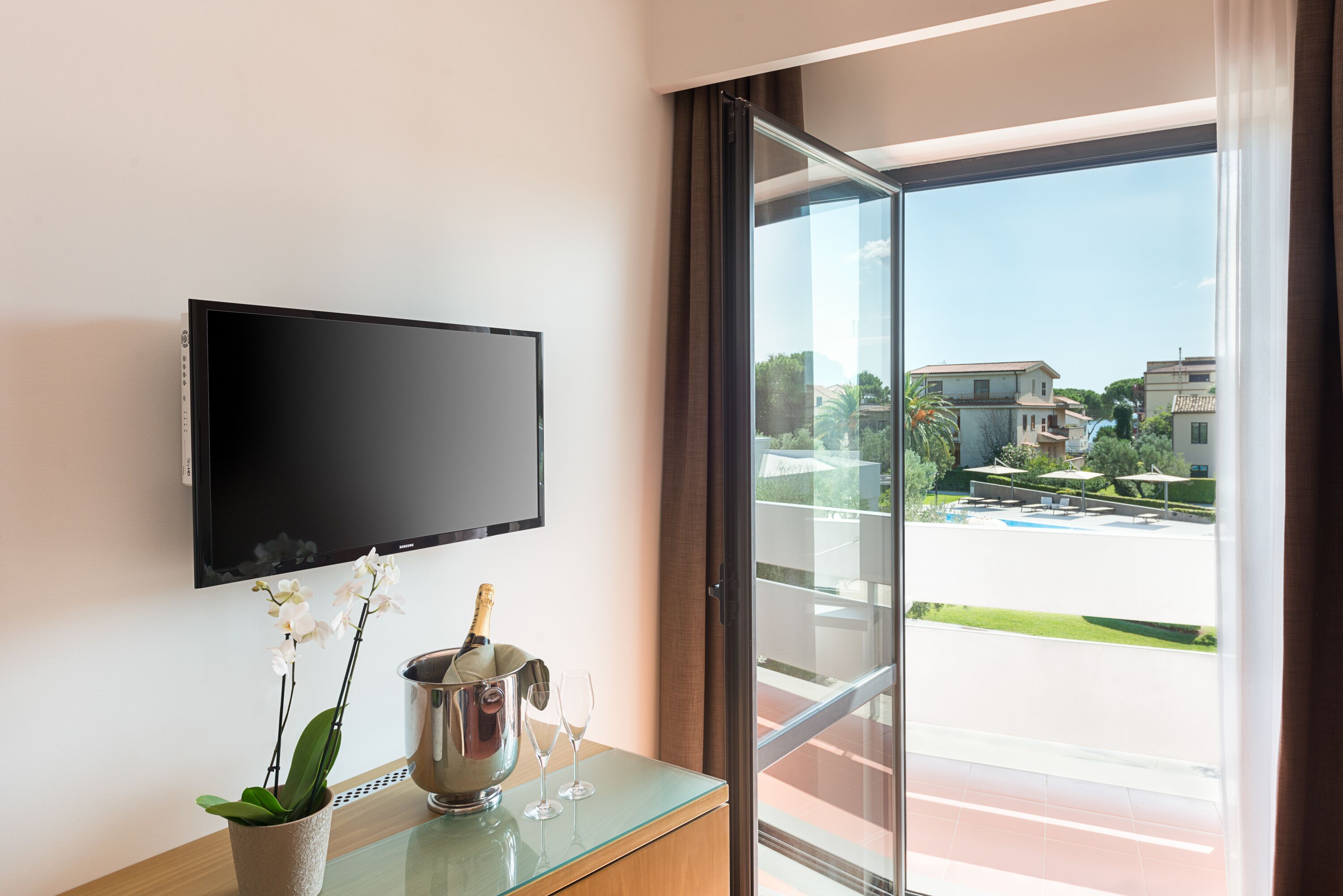 Une chambre d’hôtel moderne dotée d’un téléviseur à écran plat fixé au mur, d’une table en verre avec une bouteille de champagne et des verres, et offrant une vue sur la ville par le biais d’une grande fenêtre.