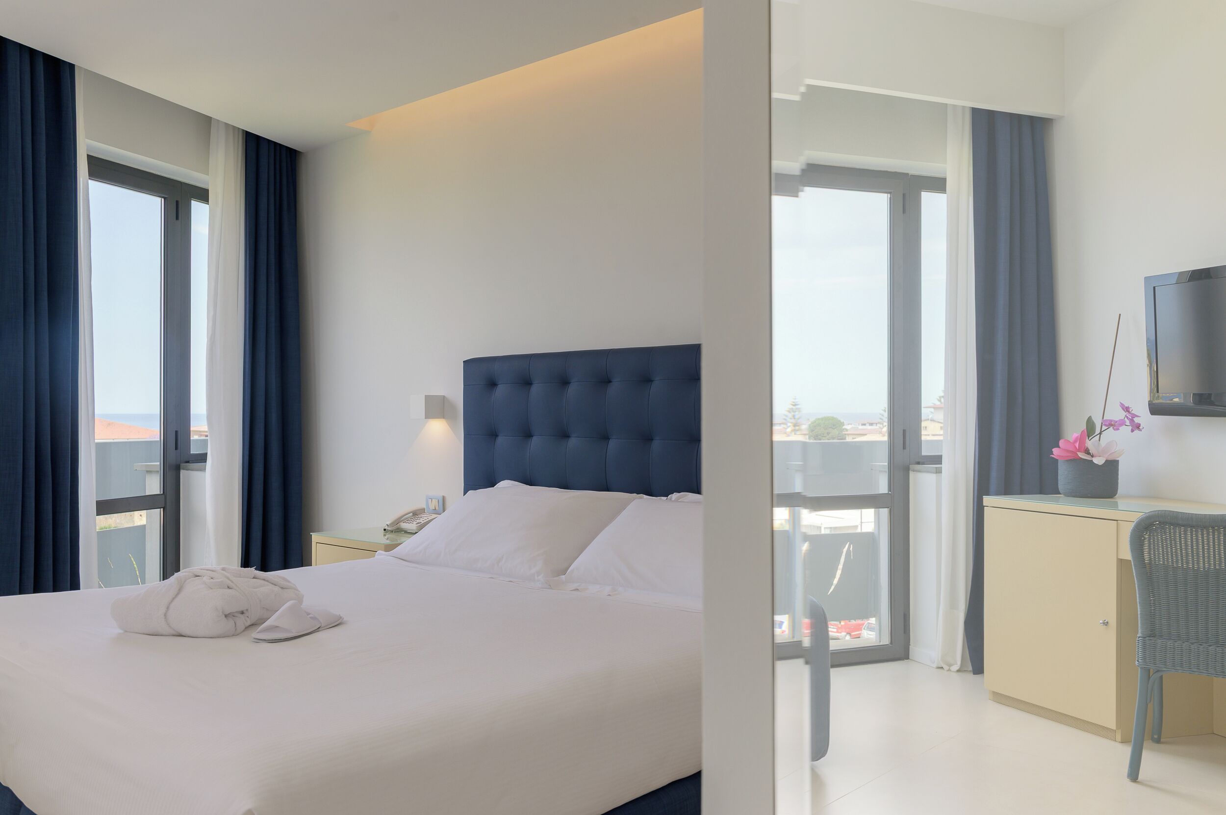 A hotel room with a bed, a desk, a chair, a TV, and a balcony with curtains.