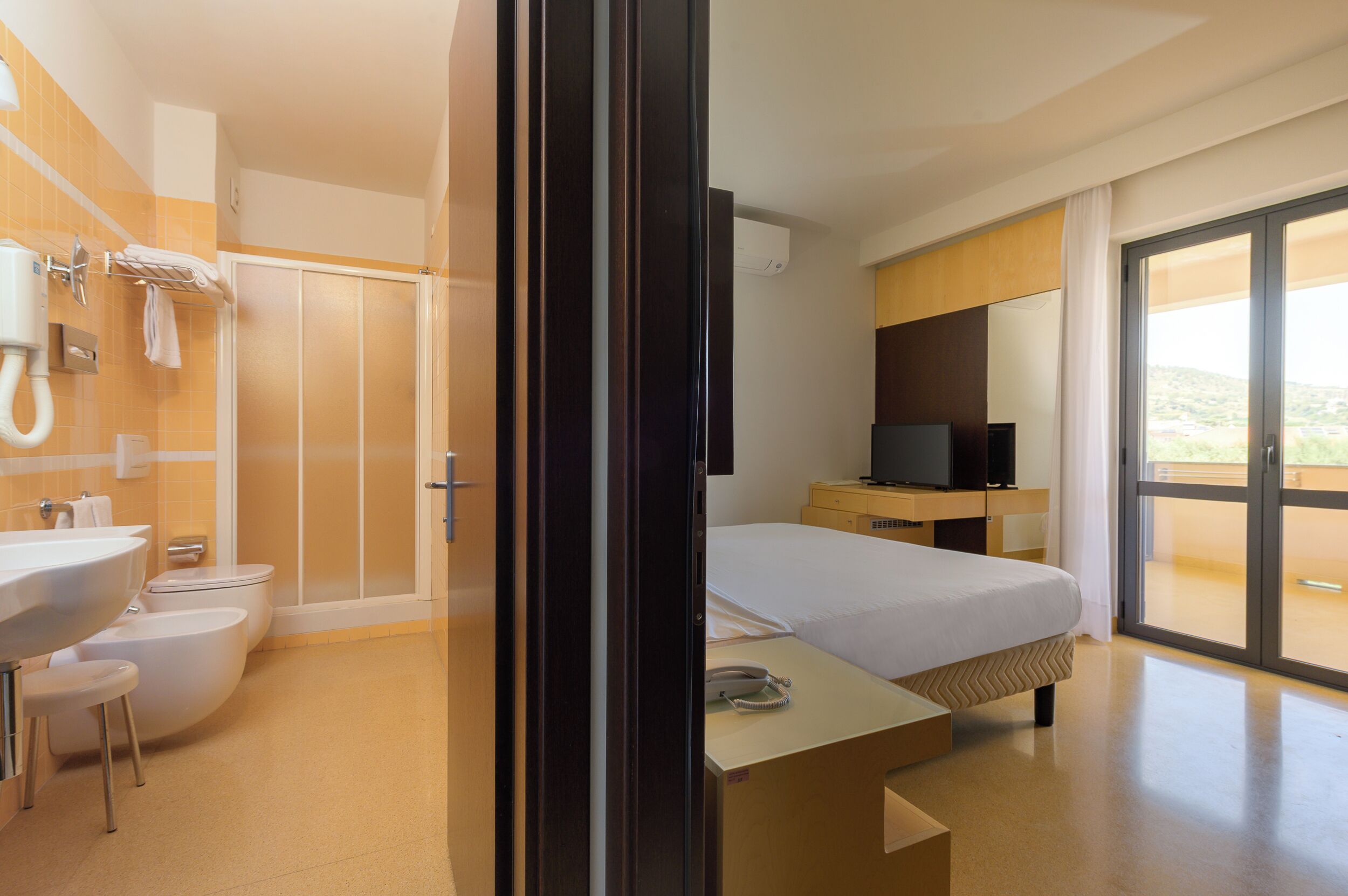 A modern hotel room with a bathroom, a bed, a TV, and a balcony with a view.