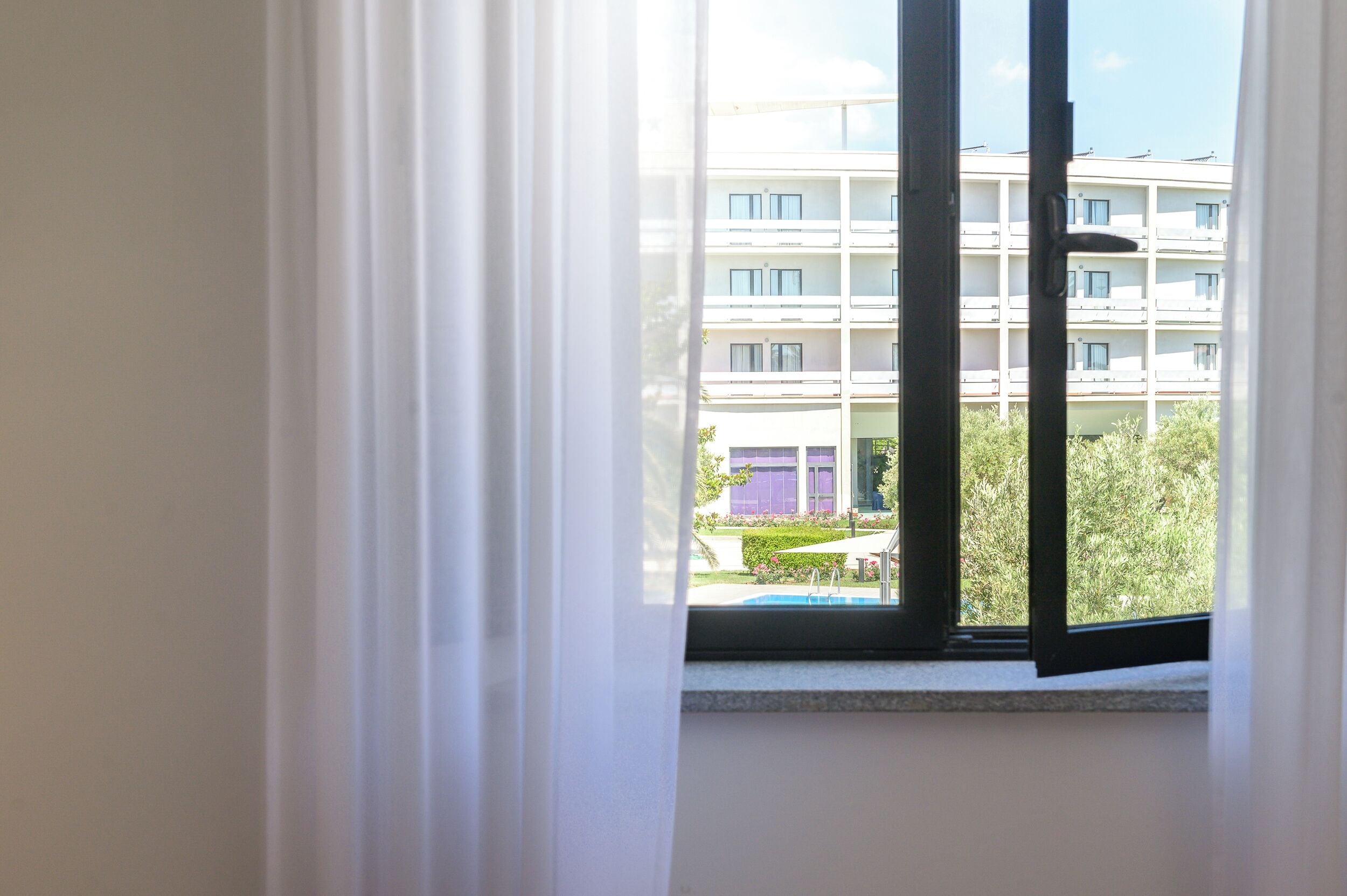 A window with white curtains, a view of a multi-story building with balconies, and a swimming pool.