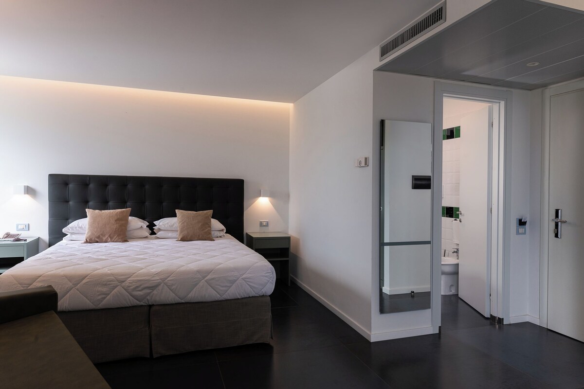A modern hotel room with a large bed, bedside tables, a mirror, and a view into the bathroom.
