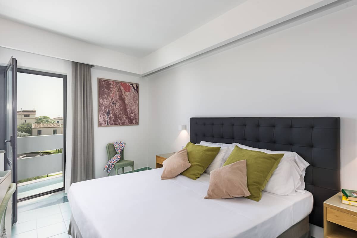 A hotel room with a large bed, a green chair, a nightstand, and a view of the outside.