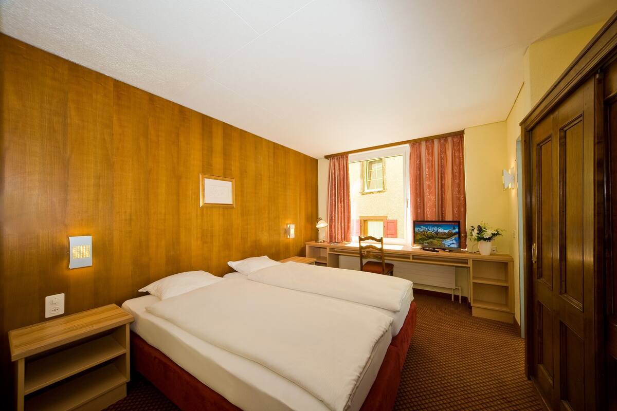 Habitación de hotel con pared de madera, cama con ropa blanca, escritorio con computadora y televisor.