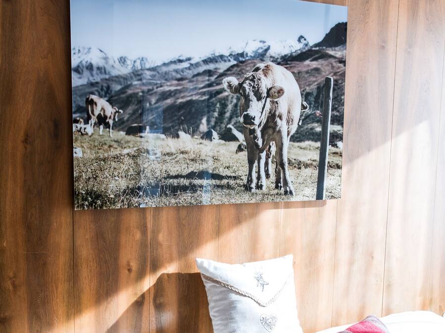 A bed with white linens and two decorative pillows, a framed photograph of a cow on a mountain, and wooden paneling on the wall.