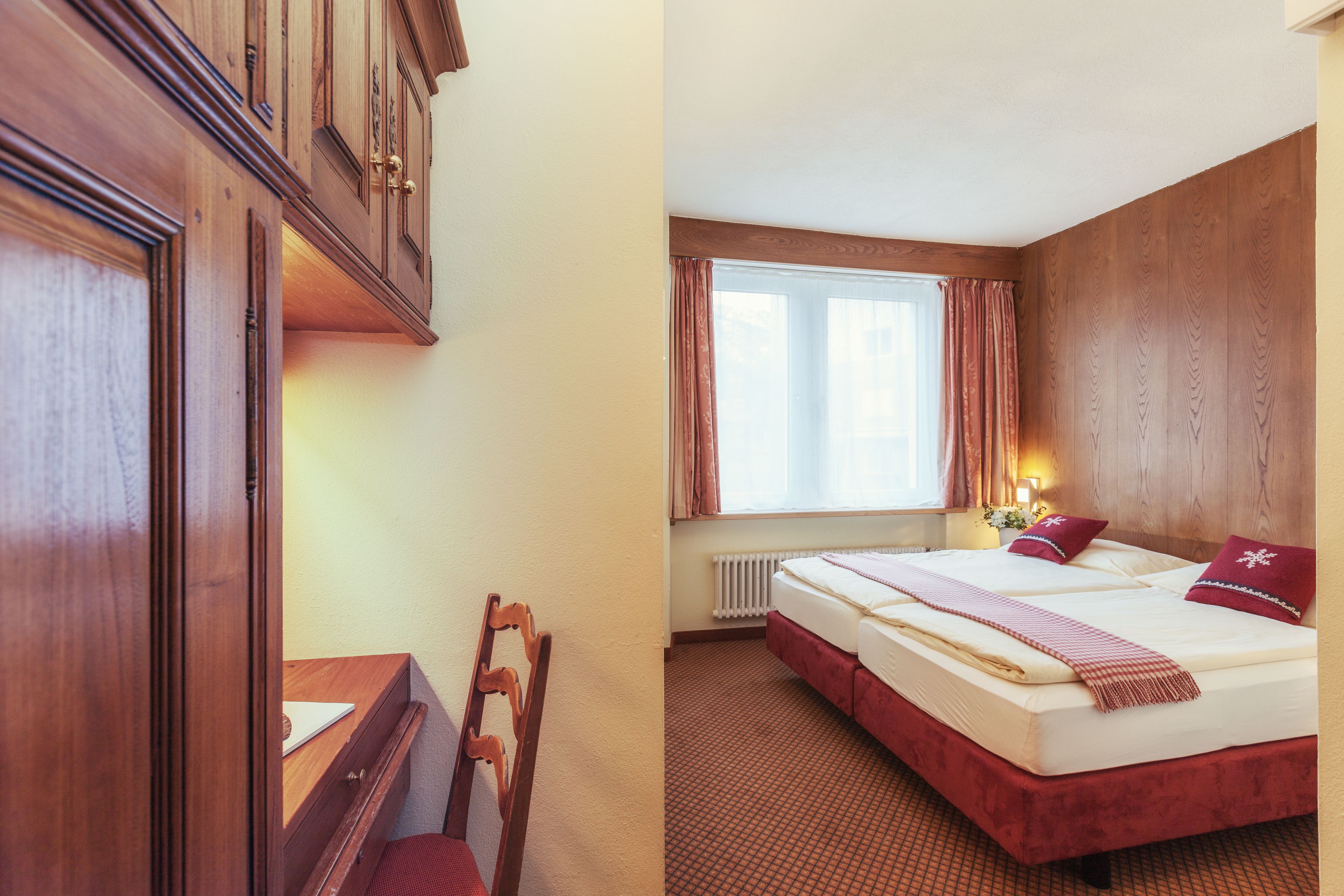 A hotel room with a wooden wardrobe, a desk with a laptop, a bed with red bedding, and a window with curtains.