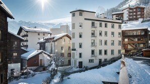 A snow-covered mountain town with a mix of traditional and modern buildings, including a prominent multi-story hotel with balconies and a ski lift visible in the background. - Le Petit CHARME-INN (Zermatt)