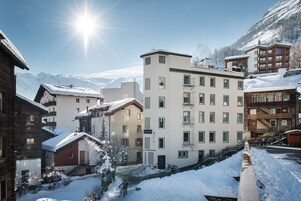 Un pueblo cubierto de nieve en la montaña con una mezcla de edificios tradicionales y modernos, incluyendo un hotel imponente de varios pisos con balcones y un telesilla visible al fondo.