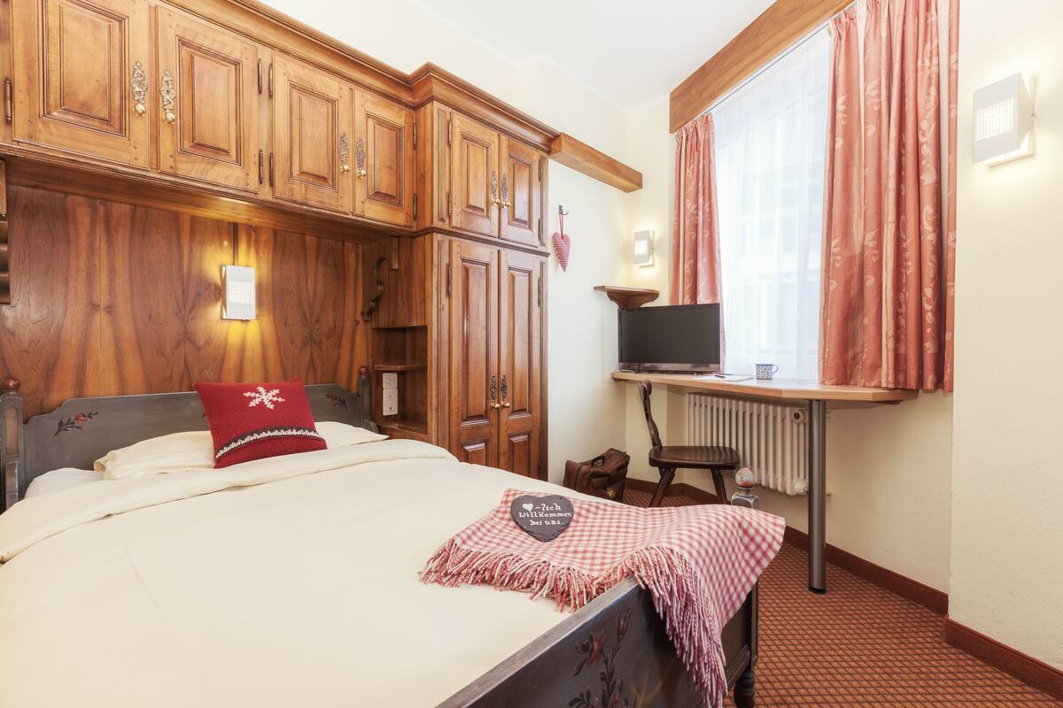 Un dormitorio con un armario de madera, una cama con almohada roja, una mesita con televisor y una ventana con cortinas.
