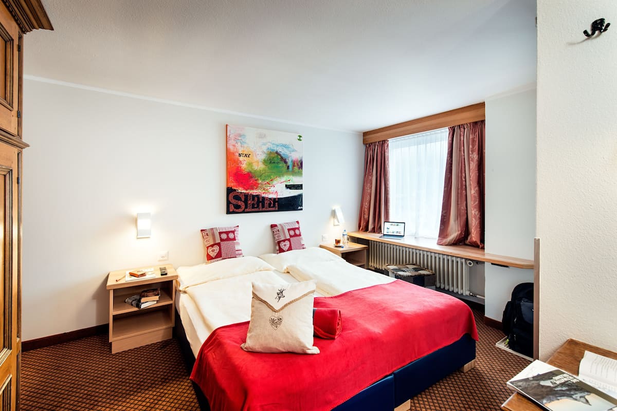 Habitación de hotel con cama, mesitas de noche, escritorio, ventana con cortinas, radiador y una obra de arte en la pared.