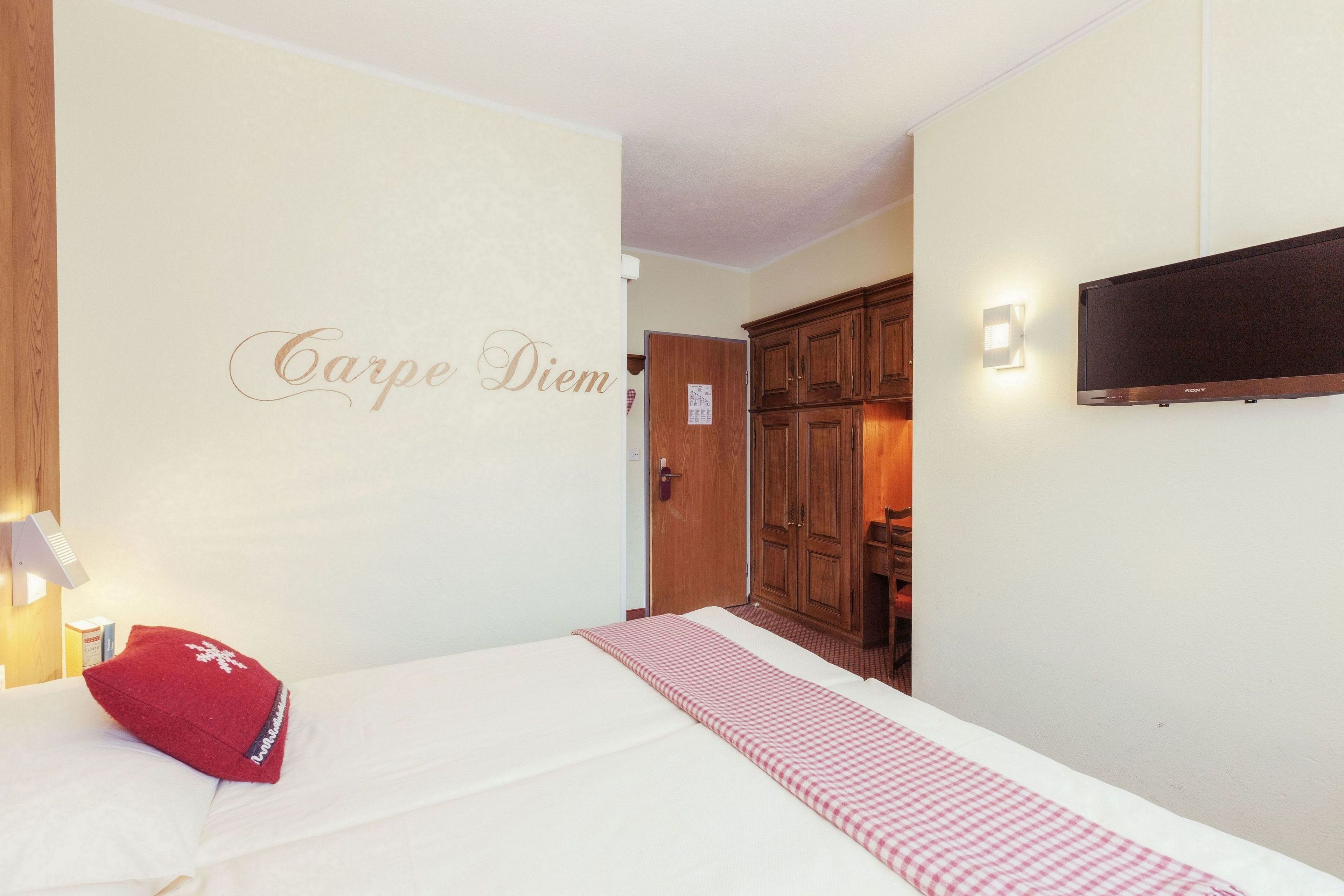 A hotel room with a bed, a red pillow, a checkered blanket, a wall-mounted TV, and a wardrobe.