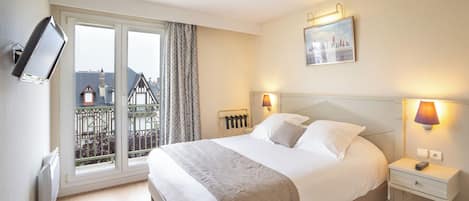 A hotel room with a large bed, a TV mounted on the wall, a balcony with a view of buildings, and a painting on the wall.