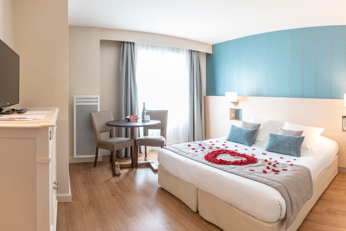 Ett hotellrum med en säng, ett skrivbord med en tv, ett litet bord med stolar och ett fönster med gardiner.