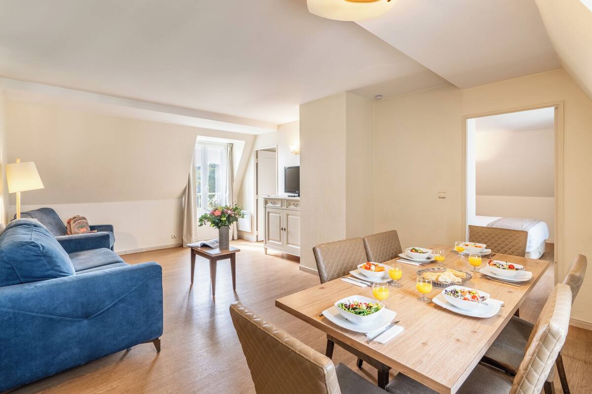 En matplats med ett träbord dukat för frukost, en blå soffa och ett kök i bakgrunden.