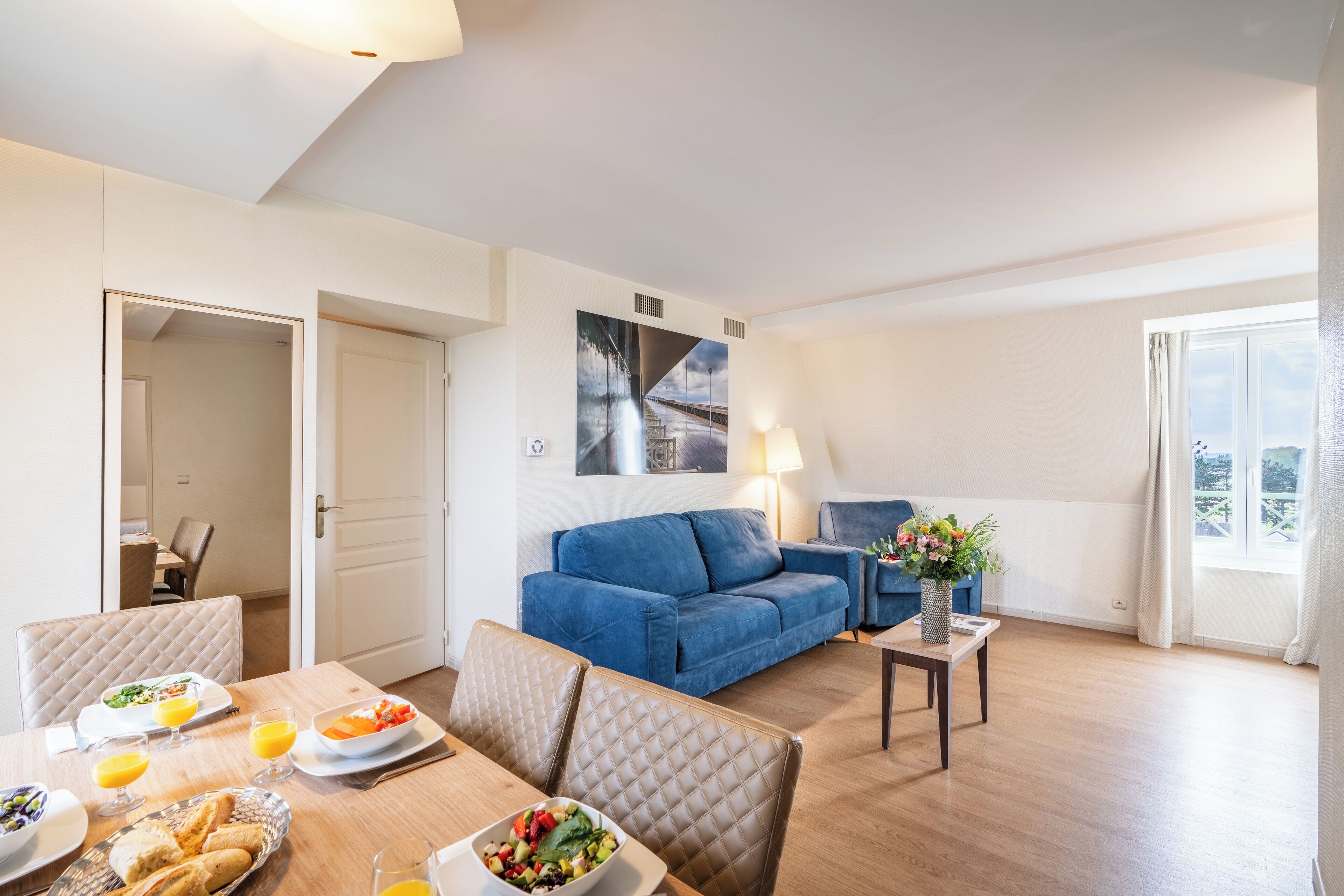 Een moderne woonkamer met een blauwe bank, een eethoek met een houten tafel en stoelen, en een groot raam met gordijnen.