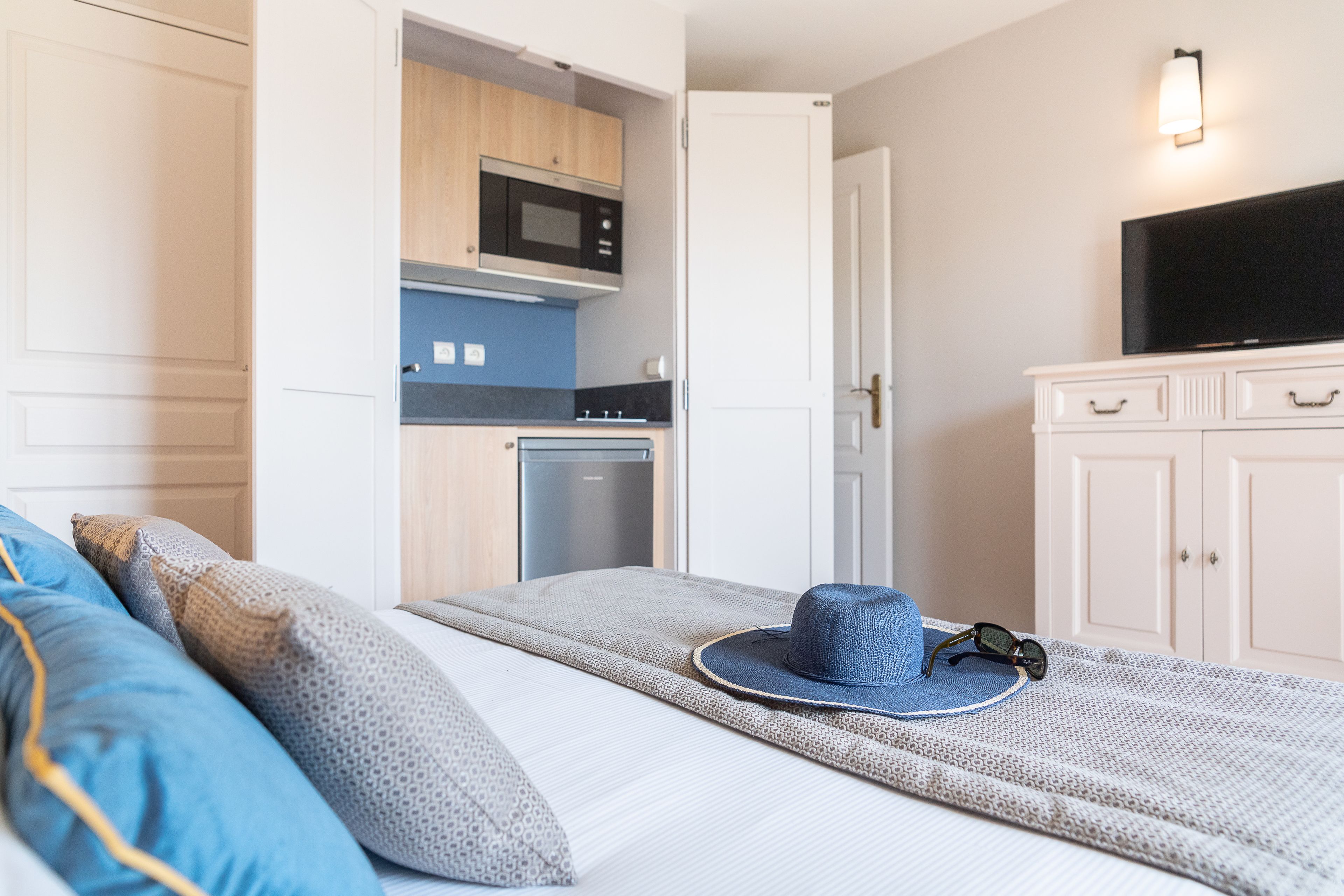 Ein Schlafzimmer mit einem Bett, Kissen, einem Hut und Sonnenbrillen auf dem Bett. Es gibt eine Küchenzeile mit einer Mikrowelle und einem Fernseher im Zimmer.