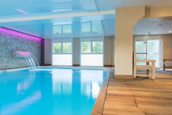 Ein Hallenschwimmbad mit klaren Glaswänden, Holzboden und Wasserfall.
