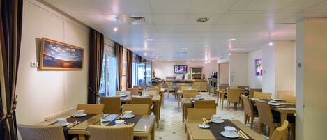 A hotel dining area with multiple tables and chairs, a television in the background, and a painting on the wall.