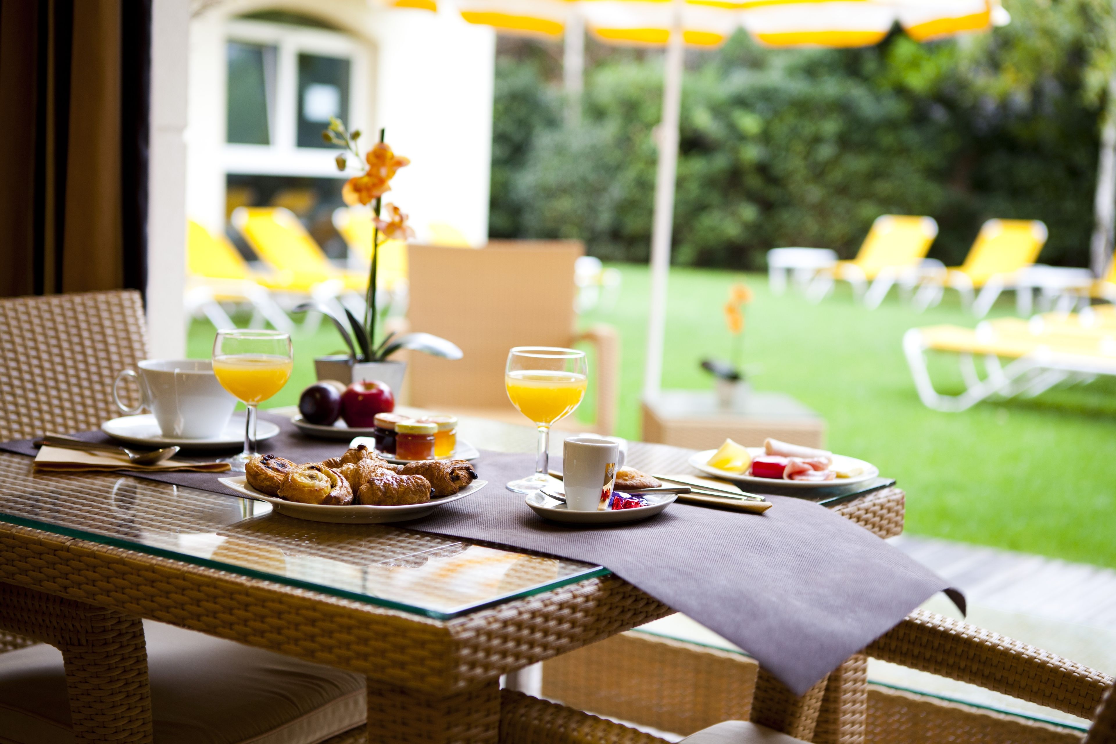 A breakfast table with croissants, fruit, and orange juice, set on a wicker table with a view of a garden and outdoor seating.