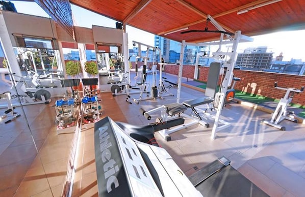 A gym with various exercise equipment, including treadmills and weight machines, under a covered area with a city view.