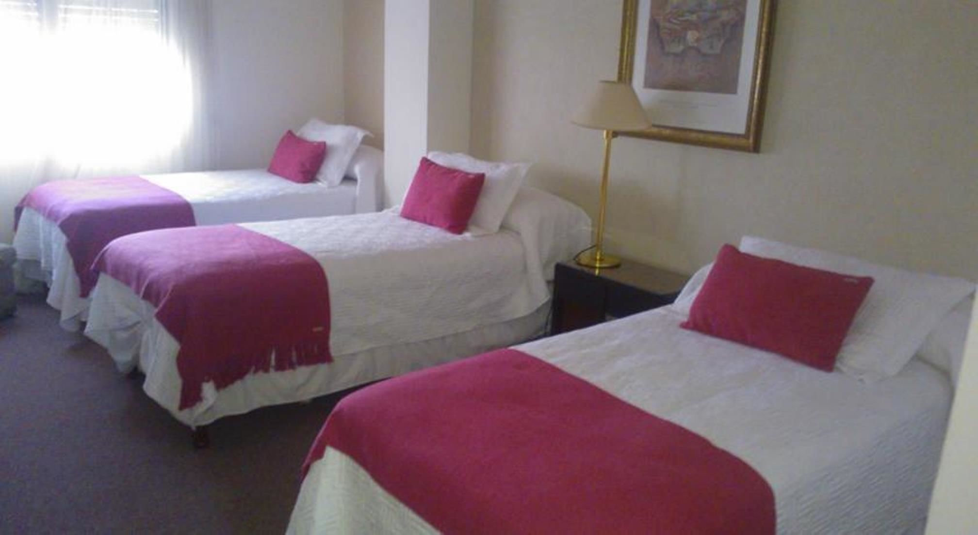 A hotel room with three single beds, each with white bedding and a red accent pillow. There is a lamp on a bedside table and a framed picture on the wall.