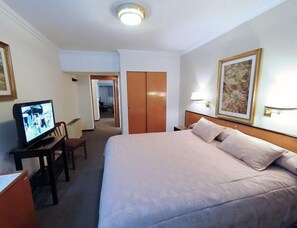 A hotel room with a bed, a desk with a television, a chair, and a door leading to another room. - Hotel Centro (Santiago del Estero)