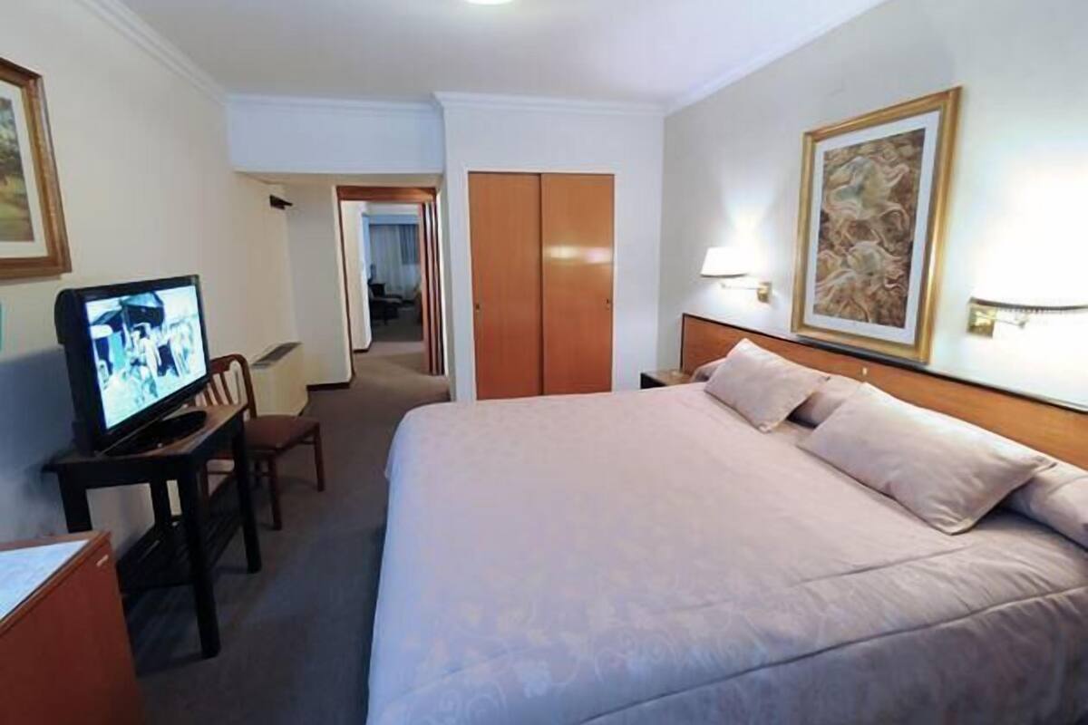 Habitación de hotel con una cama grande, un televisor en un escritorio, una puerta que conduce a otra habitación y un cuadro en la pared.