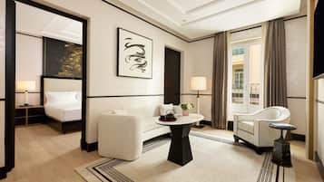Habitación de hotel con cama, sofá, mesa con florero y tetera, una silla, televisor y ventana grande con cortinas.