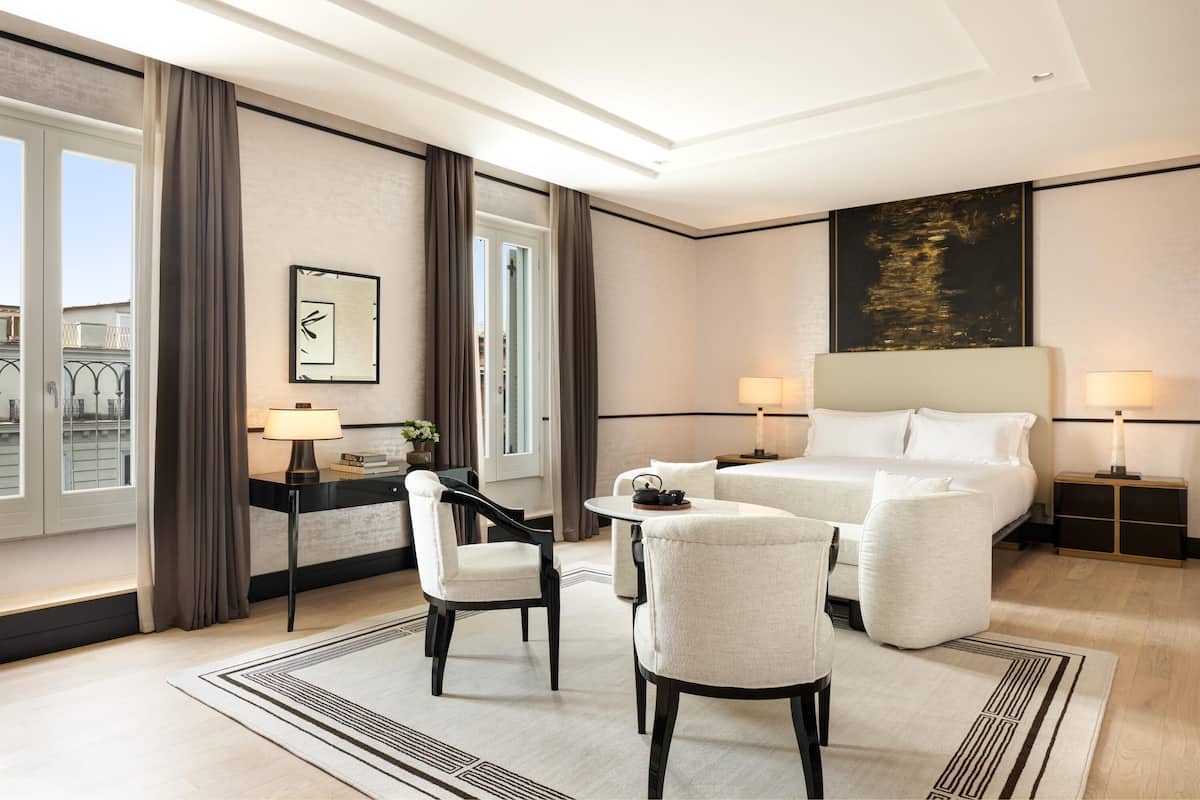 A hotel room with a large bed, two bedside tables with lamps, a desk with a chair, and a view of the city through large windows.