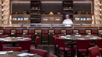 Ein Restaurant mit runden Tischen und roten Stühlen, eine Bar mit Regalen voller Flaschen und Gläsern, und ein Koch im Hintergrund.