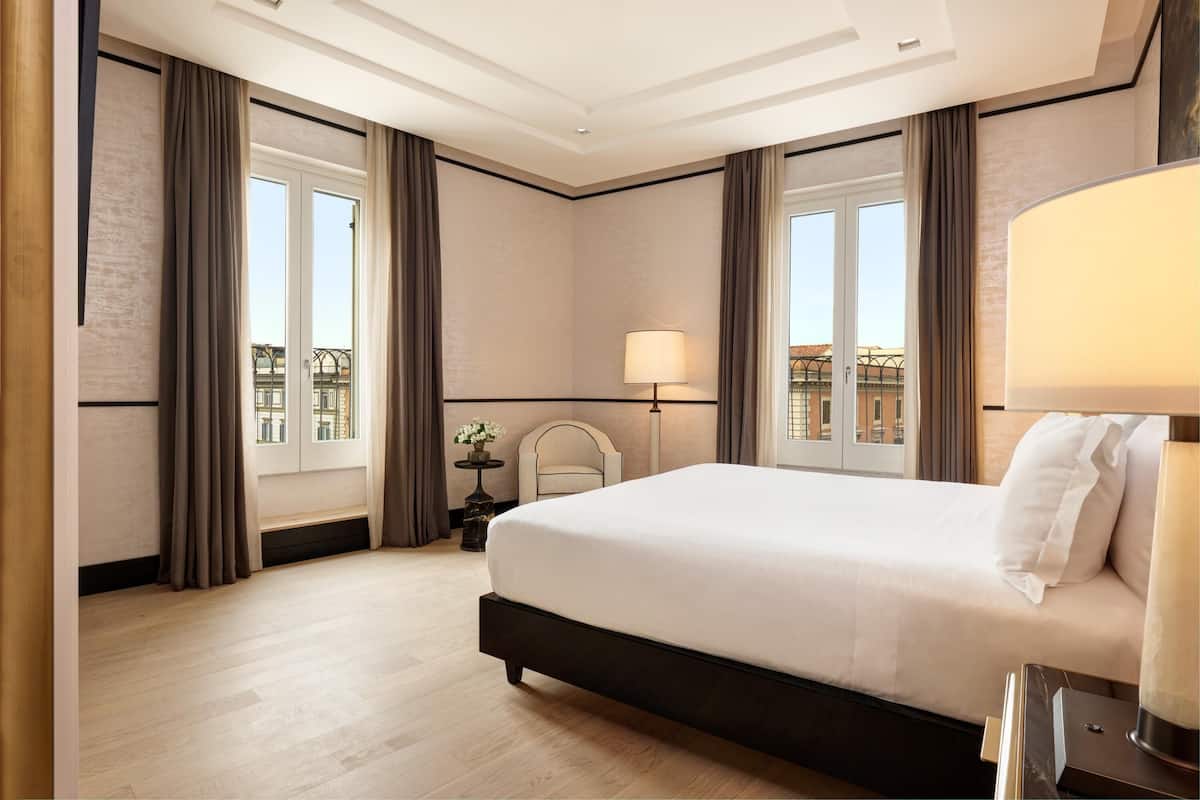 A hotel room with a large bed, a bedside table with a lamp, a small table with a vase, and a view of buildings through the window.