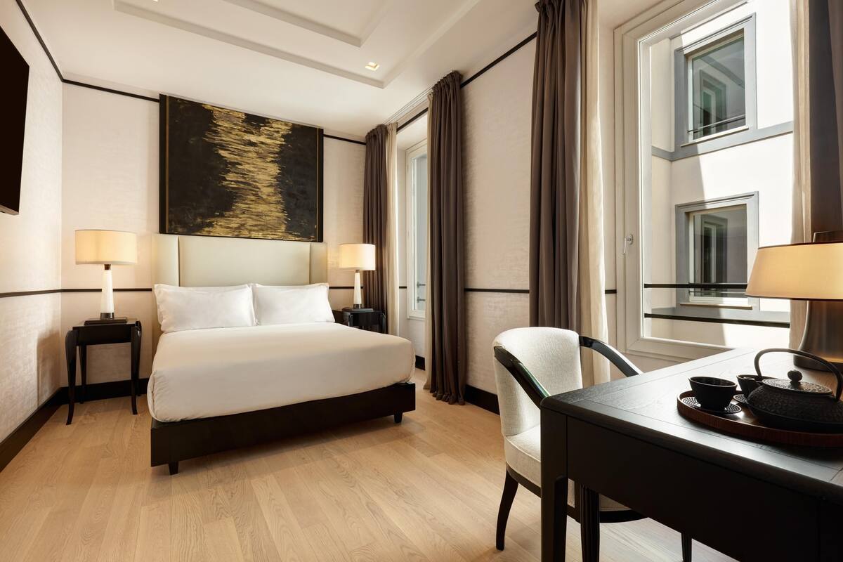 A hotel room with a large bed, two bedside tables with lamps, a chair, a table with a teapot, and a painting on the wall.