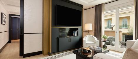 A modern hotel room with a flat-screen TV mounted on a dark cabinet, a comfortable sofa, a small table with a tray, and a view of the outside balcony.