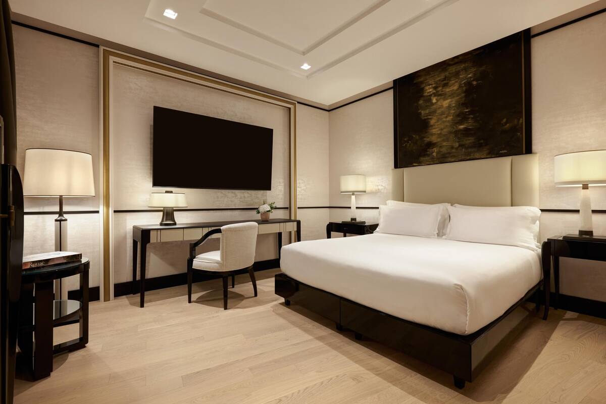 A modern hotel room with a large bed, a desk with a chair, a flat-screen TV, and two bedside lamps.