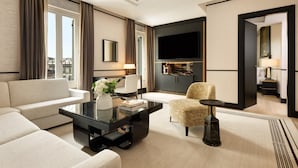 A modern hotel room with a sofa, armchair, and a glass coffee table. There is a flat-screen TV mounted on the wall and a lamp on a side table. - Nobu Hotel Roma (Rome)