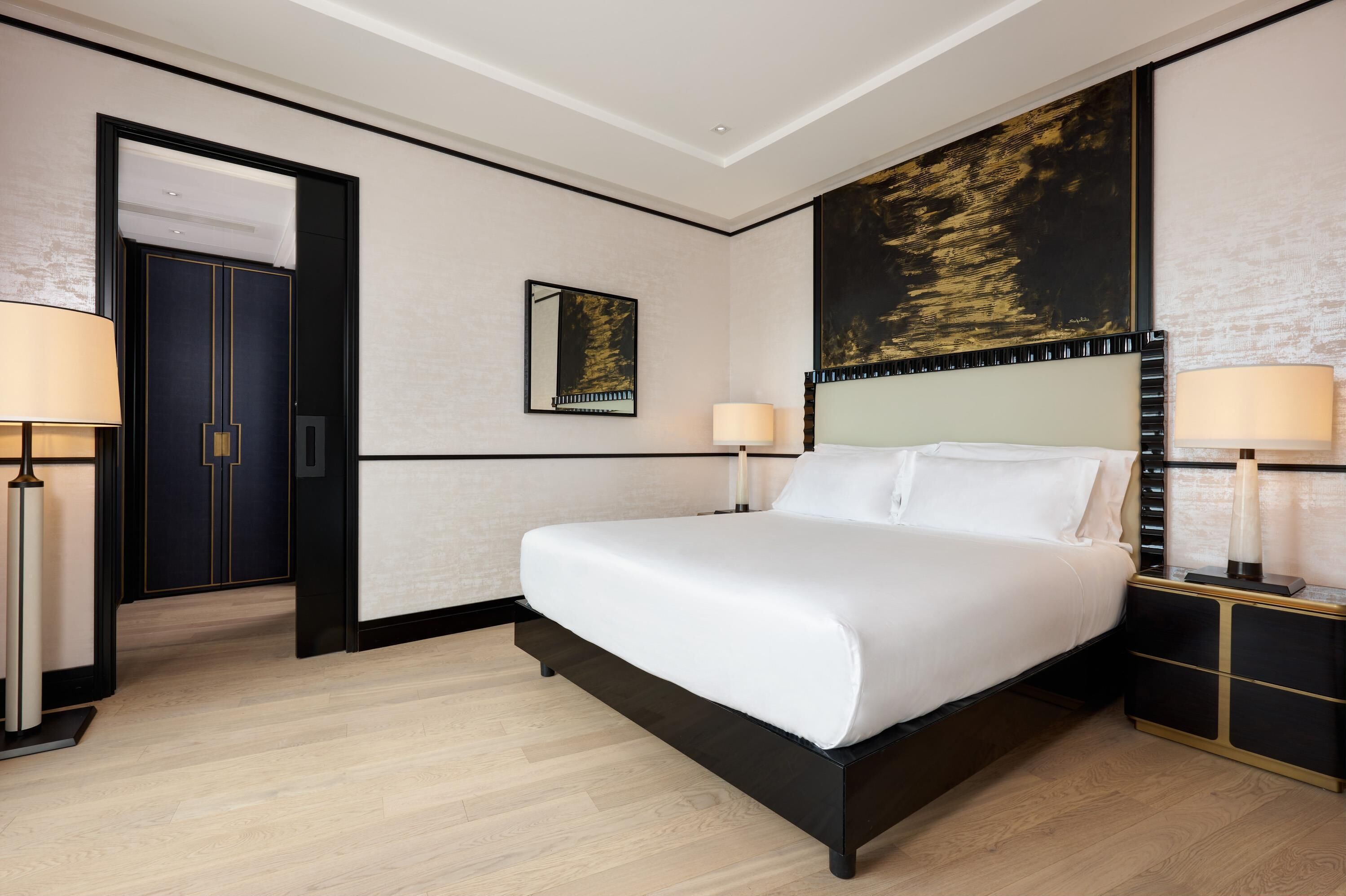 Une chambre d’hôtel moderne avec un grand lit, deux tables de chevet avec des lampes, une porte et un grand tableau abstrait accroché au mur.