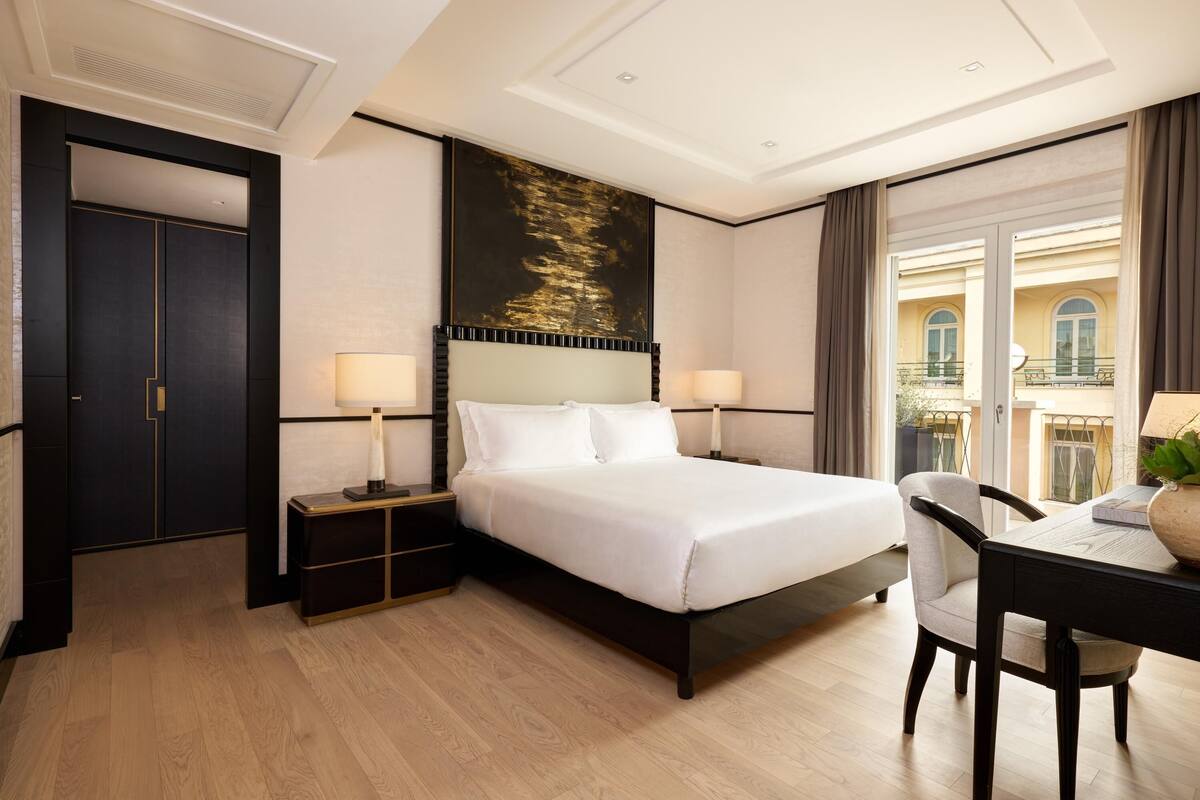 A hotel room with a large bed, two bedside tables with lamps, a desk with a chair, and a large window with curtains.