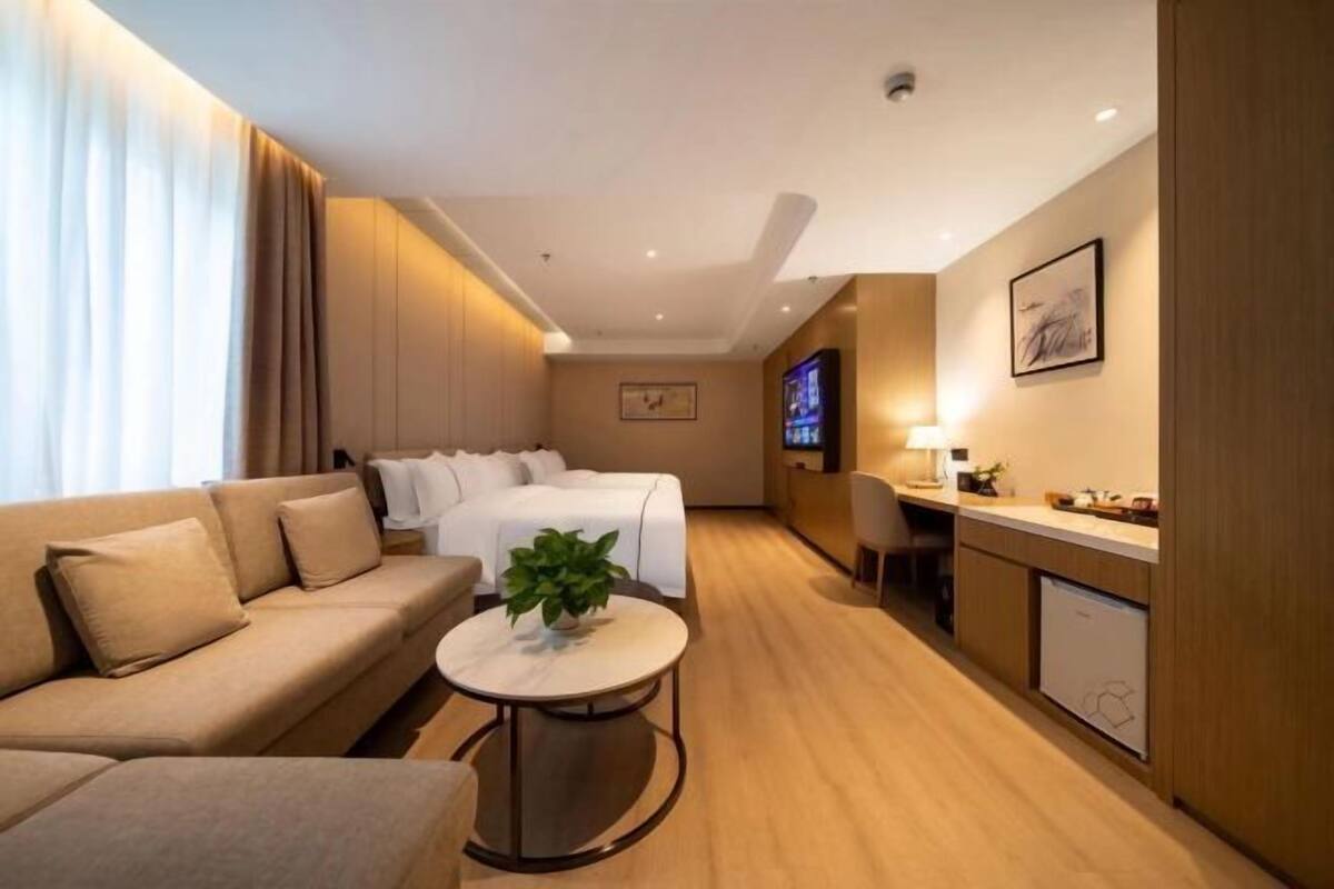 Ein Hotelzimmer mit einer Couch, einem Bett, einem kleinen Tisch mit Pflanze, einem Fernseher, einem Schreibtisch und einer Küchenzeile.