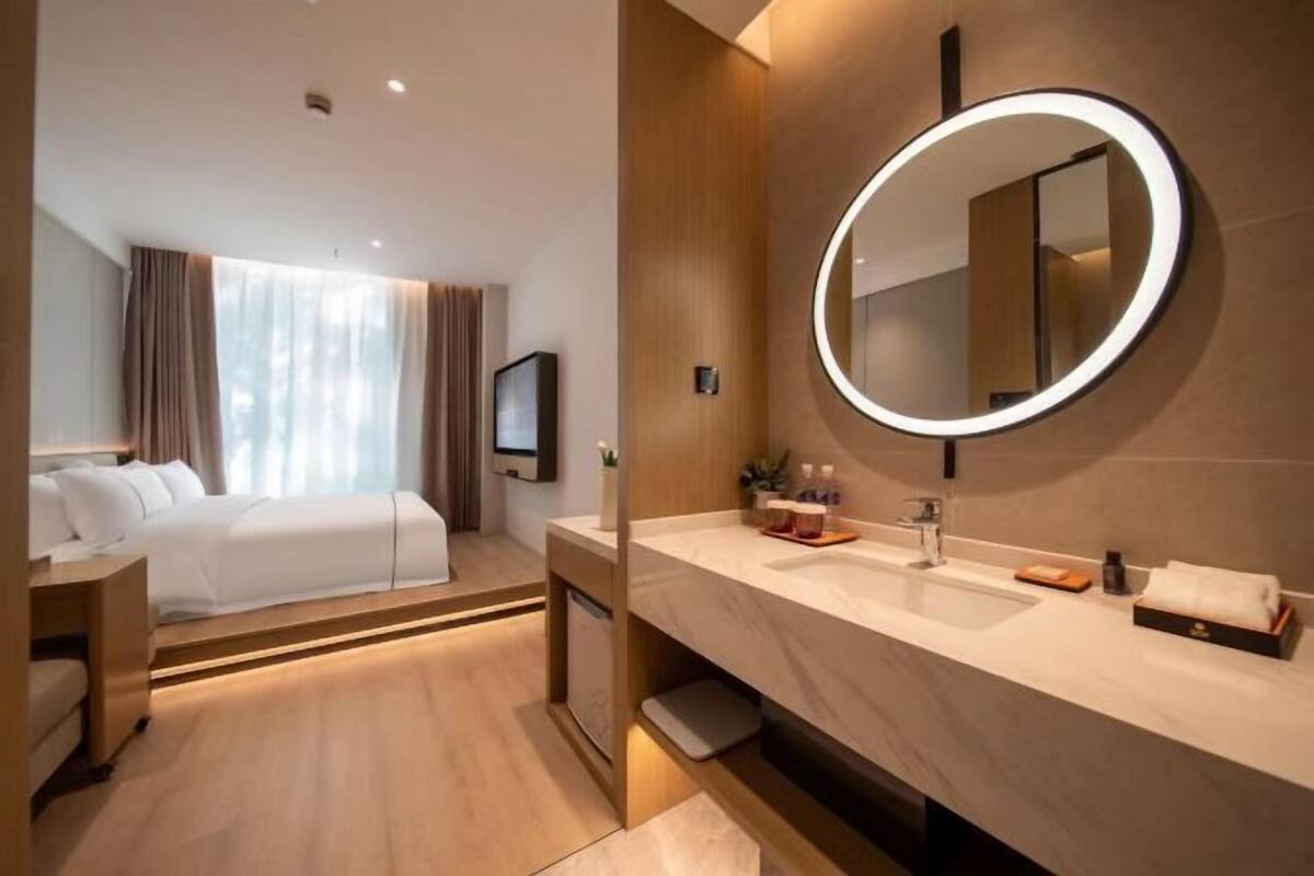 Ein modernes Hotelzimmer mit einem großen Bett, einem runden Spiegel mit Beleuchtung, einem Schminktisch mit Waschbecken und einem Fernseher an der Wand.